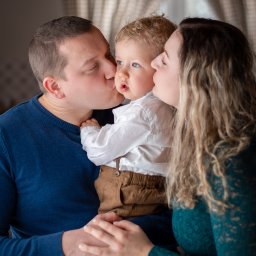 Fotoxfoto.pl Krzysztof Sobczyński - Rodzice całują w policzki swojego małego synka o jasnych włosach i niebieskich oczach, ubrany w białą koszulę i brązowe spodnie na szelkach, na tle okna z zasłonami.