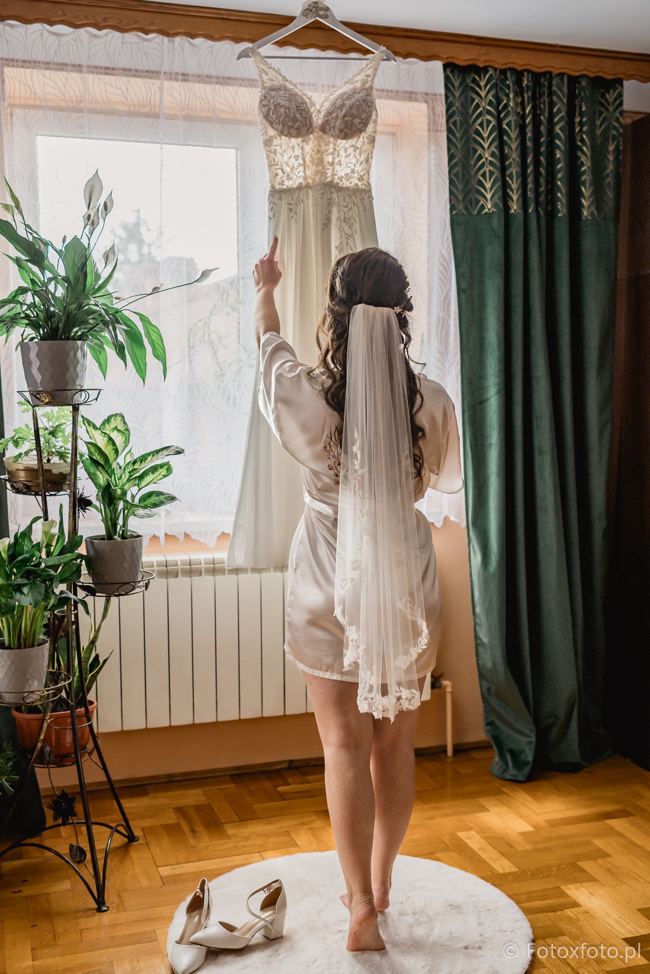 Panna młoda w szlafroku, tyłem, wskazuje na suknię ślubną wiszącą na wieszaku przed oknem. Welon na włosach, białe buty na podłodze. Dekoracje roślinne i zasłony w tle.