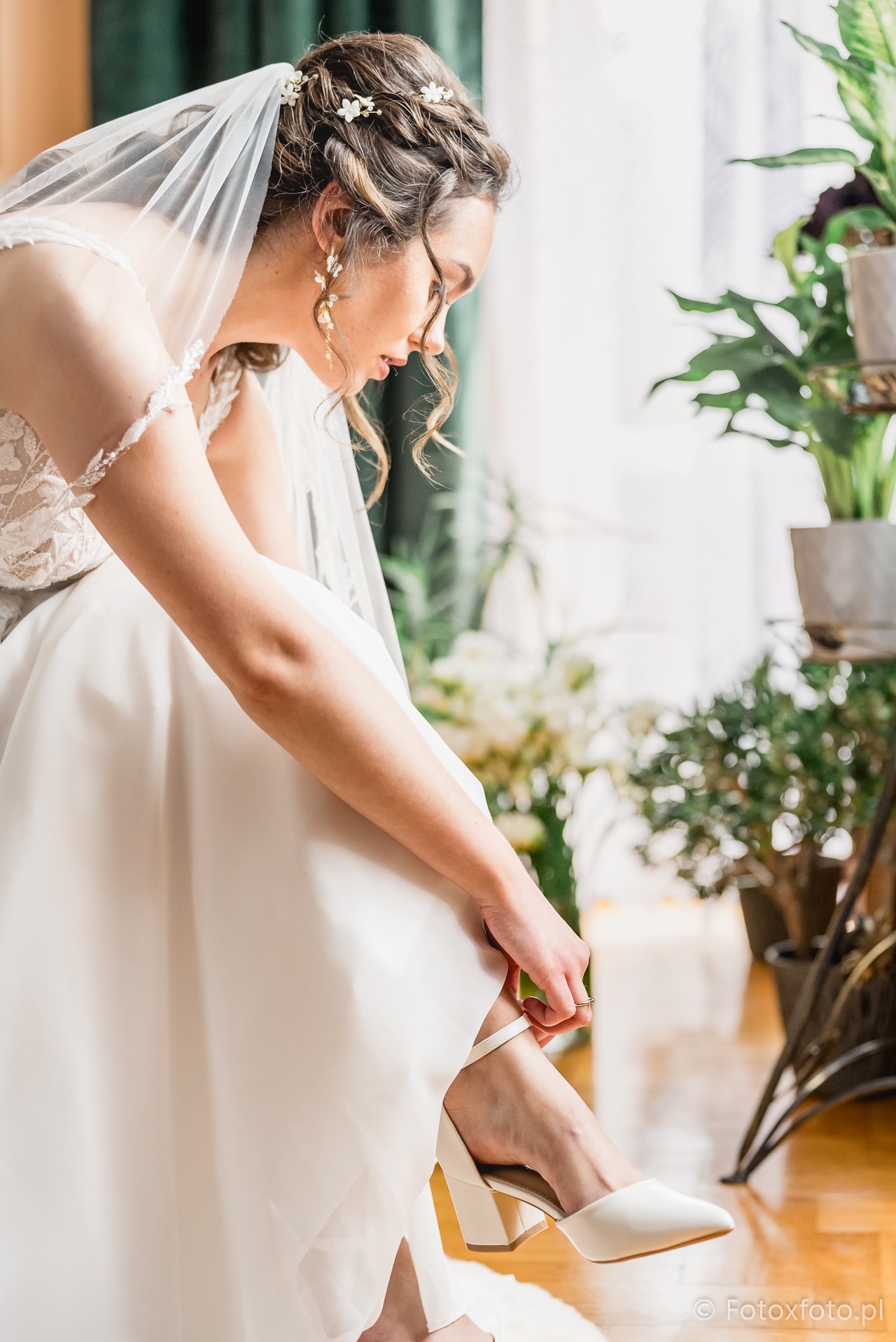 Panna młoda w koronkowej sukni, z welonem i kwiatami we włosach, zapina białe buty na tle roślin doniczkowych i jasnego okna. Delikatne światło podkreśla detale.