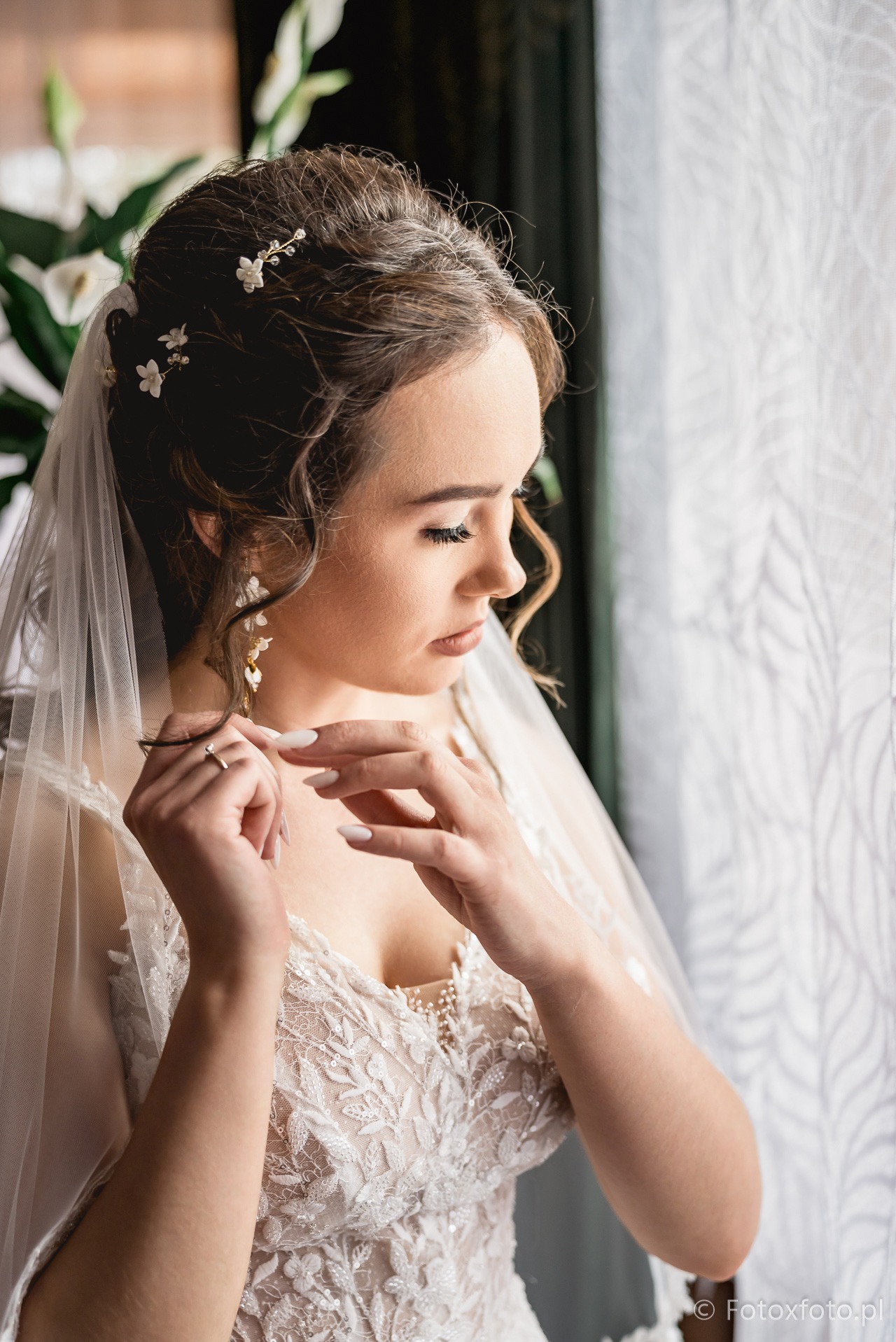 Panna młoda w koronkowej sukni i welonie, z delikatnymi kwiatami we włosach, spogląda w dół, dotykając pierścionka. Jasne, naturalne światło z okna.