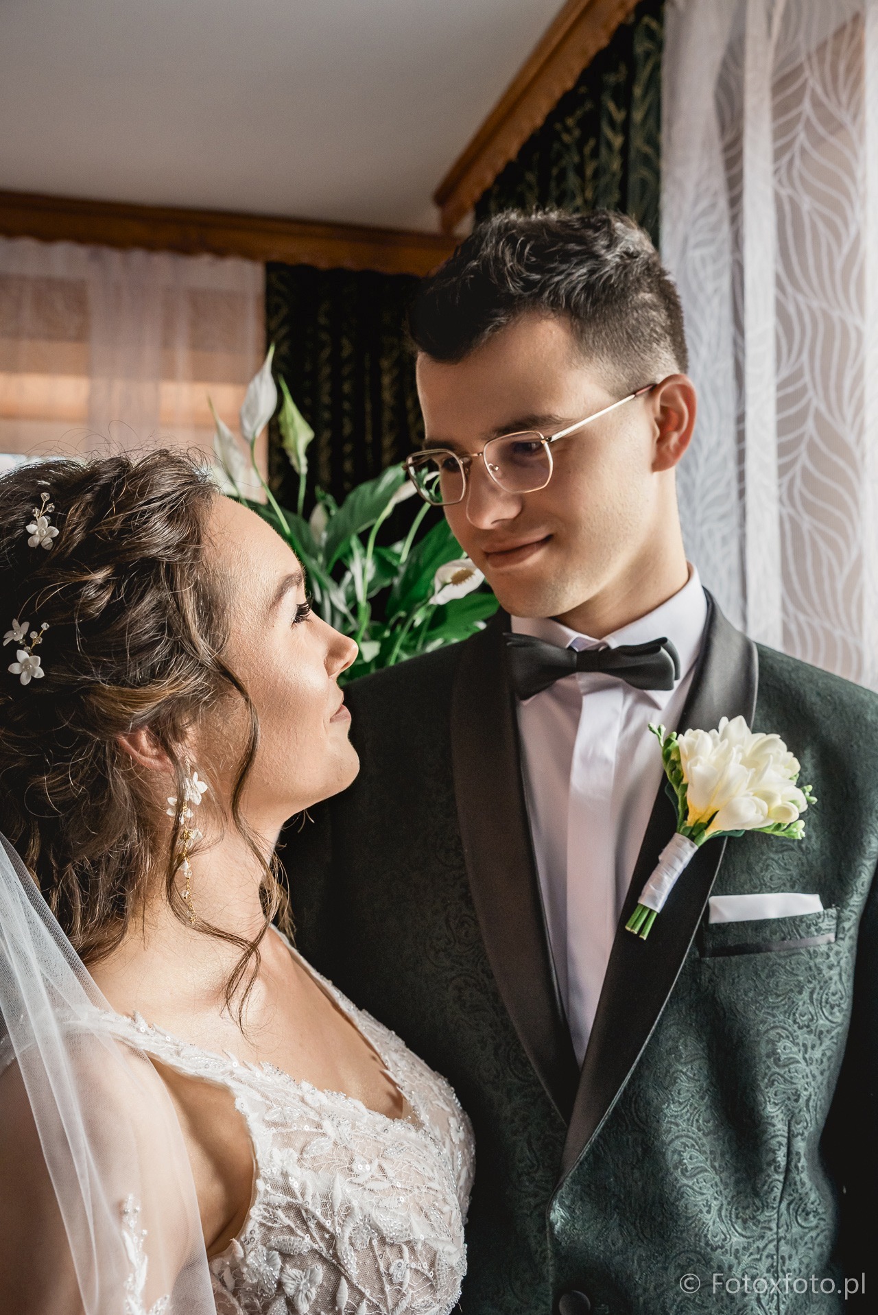 Uśmiechnięta para młoda w eleganckich strojach patrzy na siebie, on w okularach i z butonierką, ona w sukni z koronką i welonem, subtelne oświetlenie.