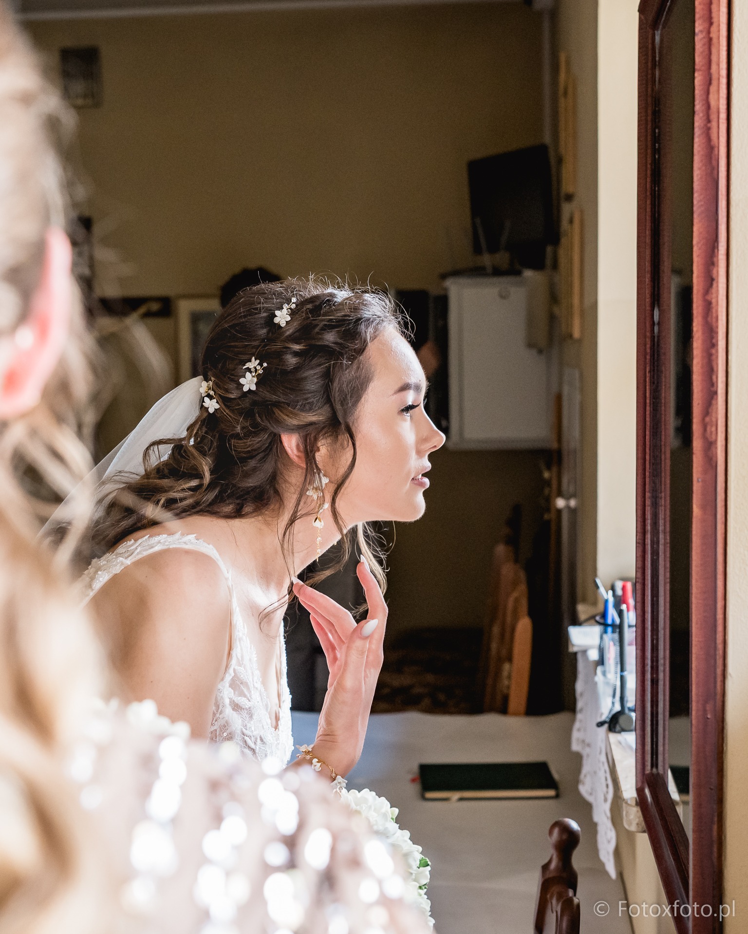 Panna młoda w białej sukni z welonem, przegląda się w lustrze, poprawiając włosy z kwiatami. Widok z profilu, delikatne oświetlenie, fragment twarzy osoby towarzyszącej.