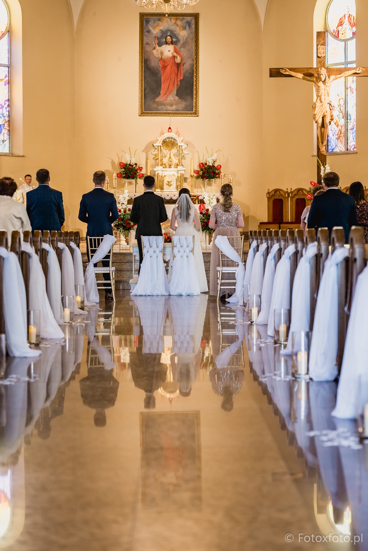 Para młoda podczas ceremonii ślubnej w kościele. Widok z tyłu na parę, gości i ołtarz. Dekoracje kwiatowe i świece. Podłoga z odbiciem.
