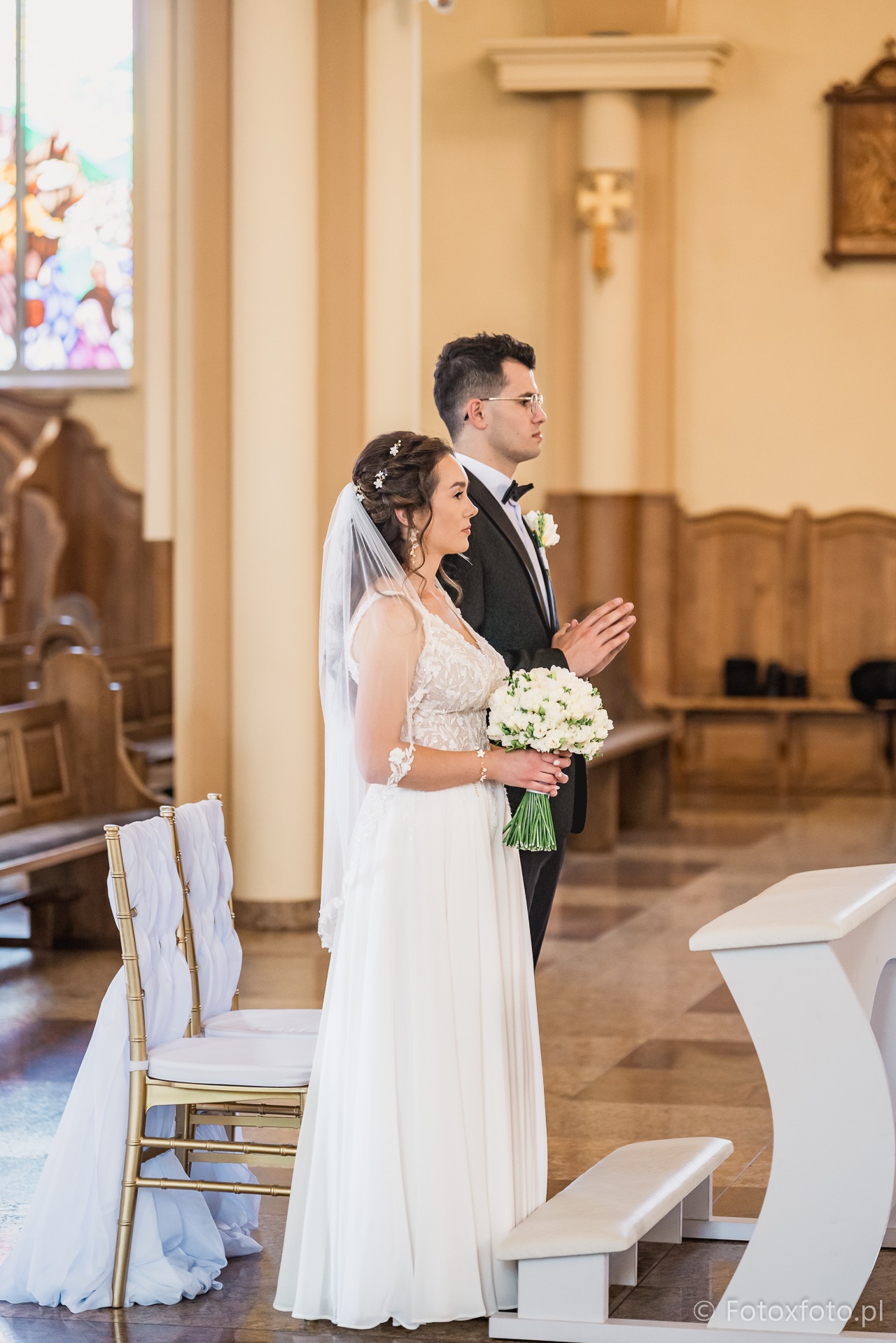 Para młoda w kościele podczas ceremonii ślubnej; panna młoda trzyma bukiet białych kwiatów, obok klęcznik i krzesła z dekoracjami.