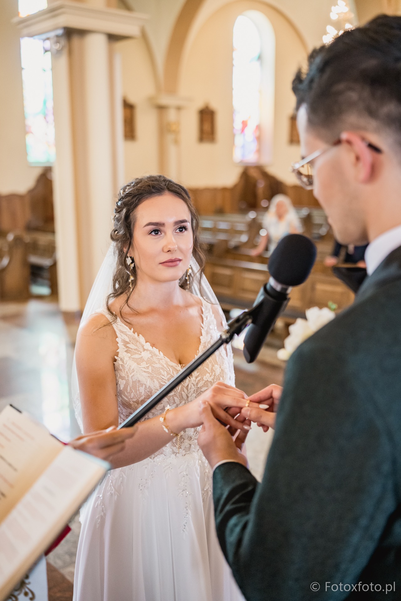 Wymiana obrączek podczas ceremonii ślubnej w kościele. Panna młoda w koronkowej sukni, pan młody zakłada jej obrączkę. W tle witraże i ławki.