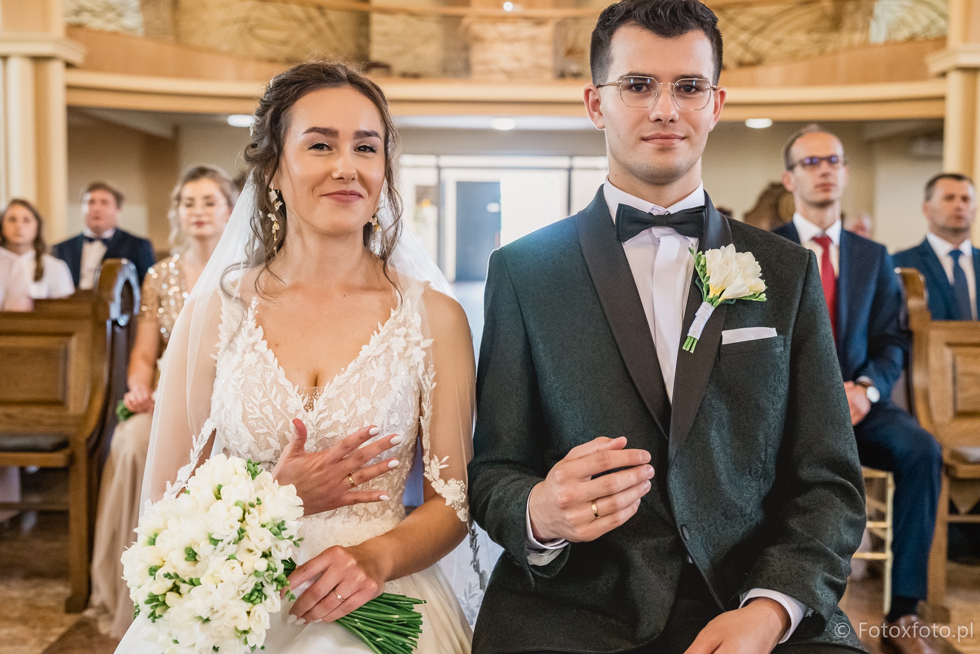 Para młoda w kościele: panna młoda w koronkowej sukni z bukietem, pan młody w ciemnozielonym smokingu z muchą. Goście w tle, drewniane ławki.