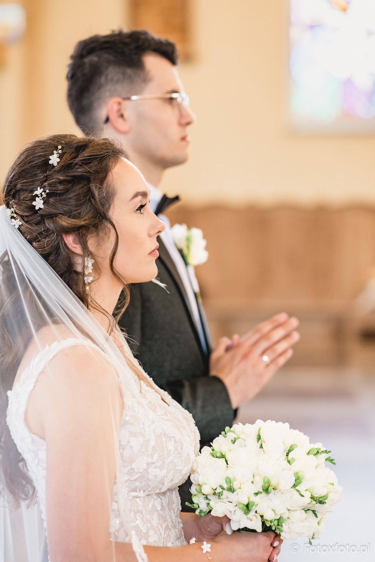 Panna młoda z bukietem frezji i pan młody w kościele podczas ceremonii ślubnej. Profil, detale sukni ślubnej i fryzury, złożone dłonie pana młodego.