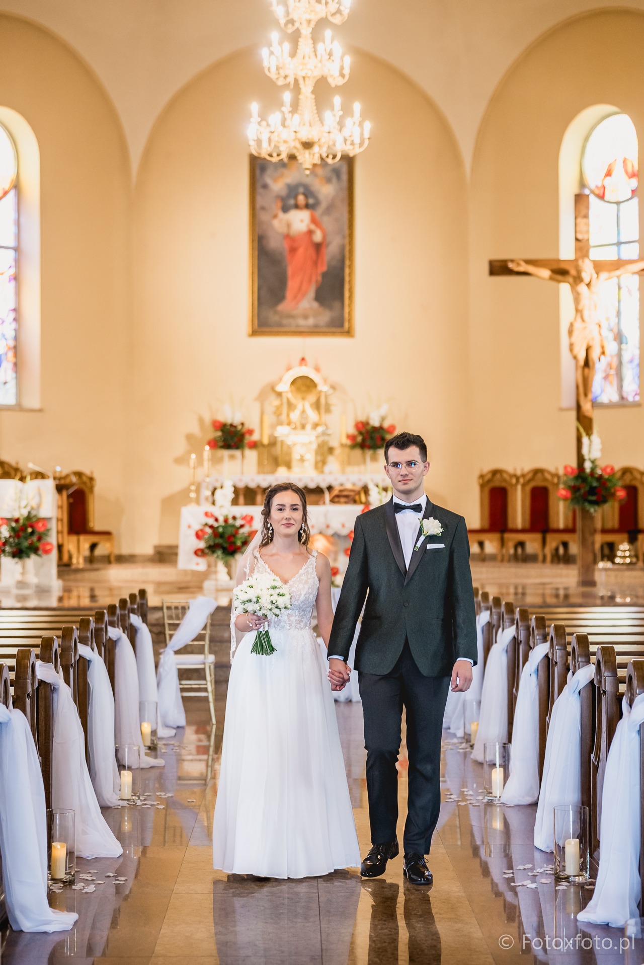 Para młoda, trzymając się za ręce, idzie w kierunku ołtarza w kościele. Dekoracje z białych tkanin i świec wzdłuż ławek. Panna młoda z bukietem.