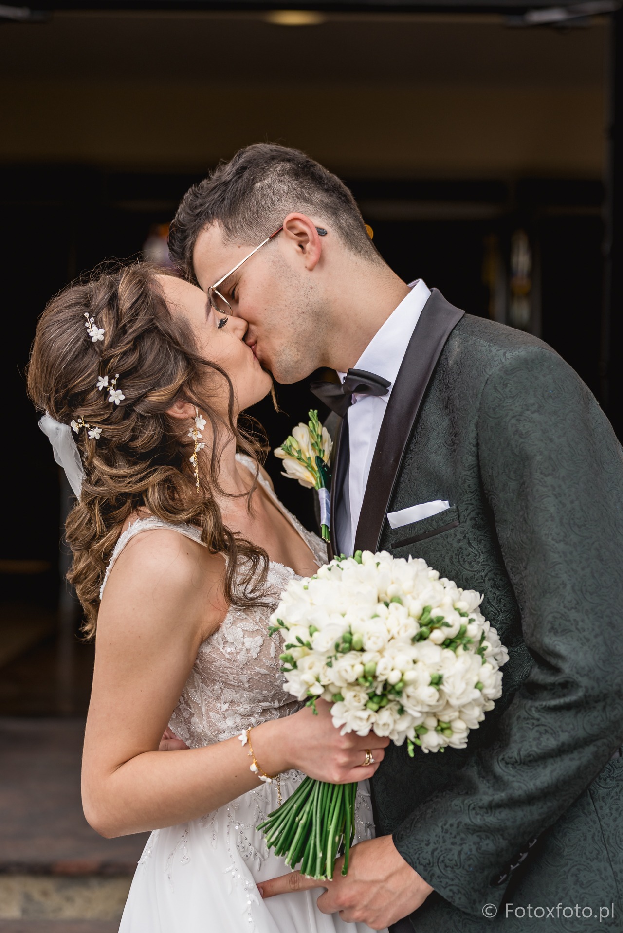 Pocałunek nowożeńców w ciemnym garniturze i sukni z bukietem frezji na tle ciemnych drzwi. Kwiaty we włosach panny młodej i okulary pana młodego.