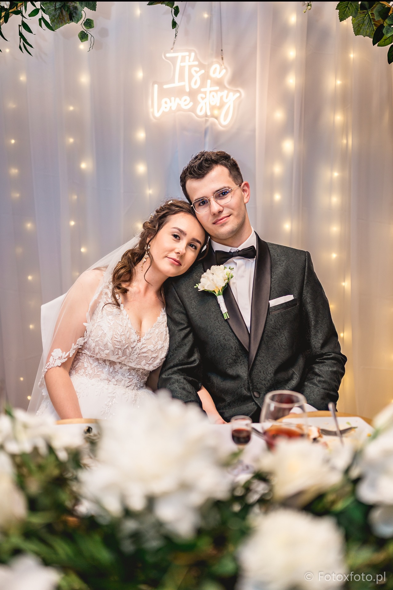Para młoda w eleganckich strojach siedzi przy stole weselnym z kwiatami i napisem neonowym 'It's a love story' w tle, uśmiechnięci, w bliskim ujęciu.
