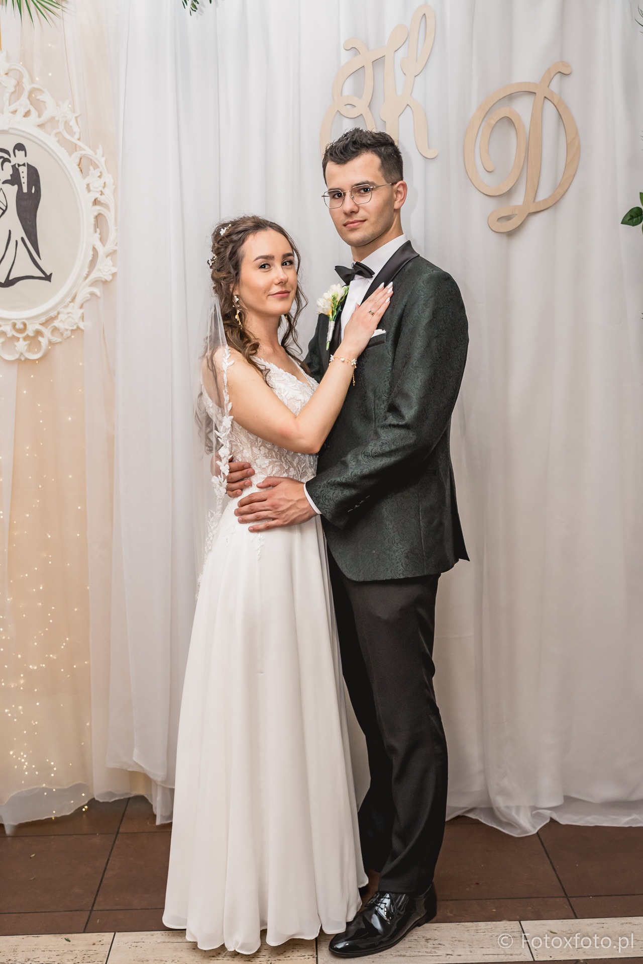 Elegancka para młoda w bliskim uścisku na tle dekoracji ślubnych. Panna młoda w koronkowej sukni, pan młody w zielonym smokingu. Romantyczna atmosfera, subtelne oświetlenie.