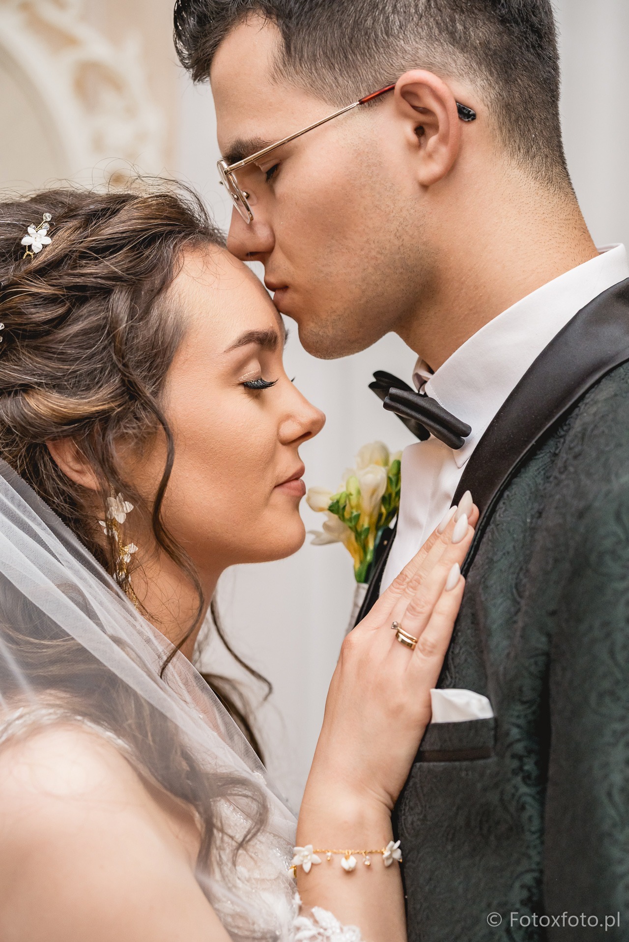 Delikatne zbliżenie: pan młody w okularach całuje pannę młodą w czoło, ona z zamkniętymi oczami, welon i kwiaty we włosach, bukiet w butonierce, profil.