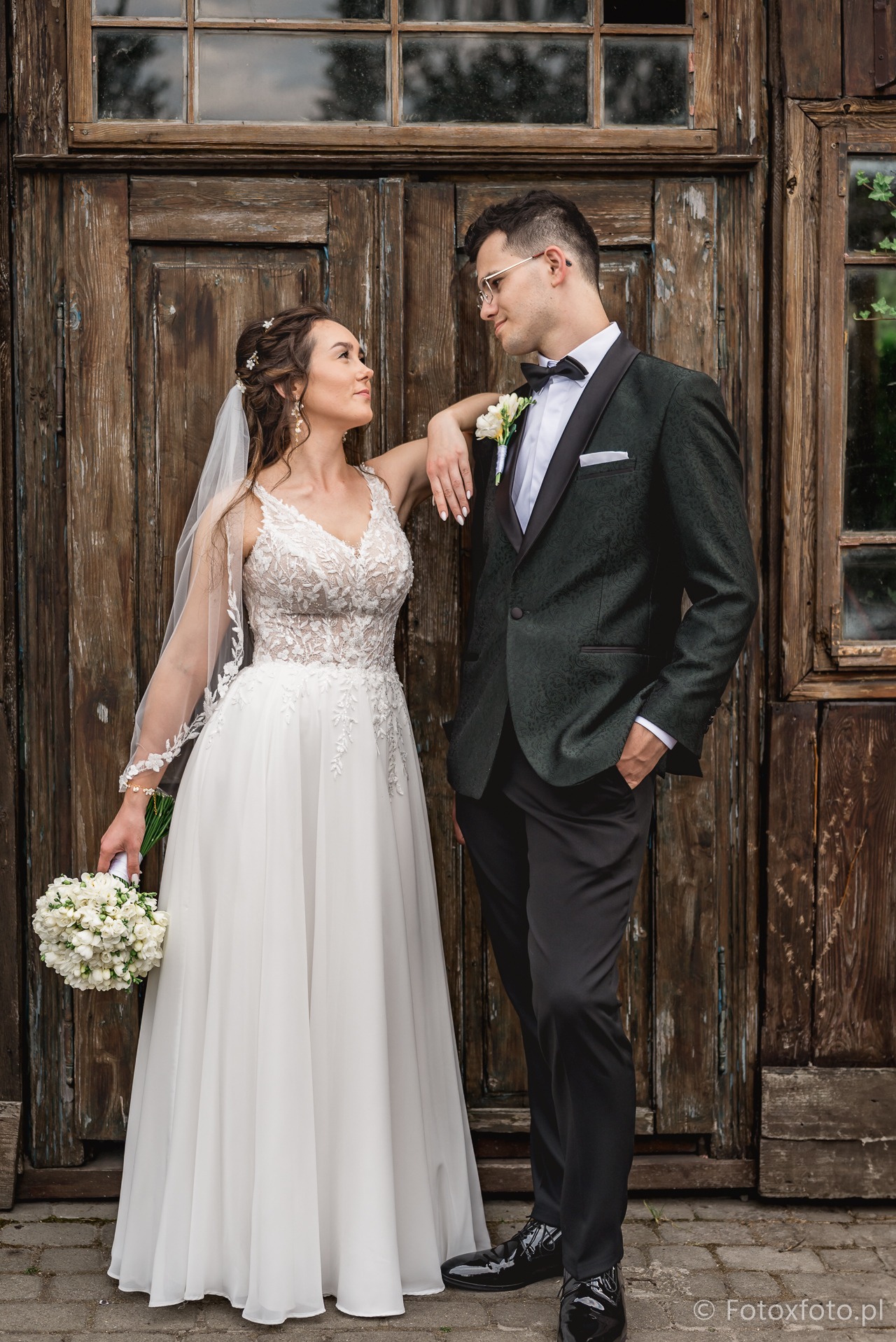 Elegancka para młoda: panna młoda w koronkowej sukni opiera się o pana młodego w ciemnozielonym smokingu na tle drewnianych drzwi z oknem.
