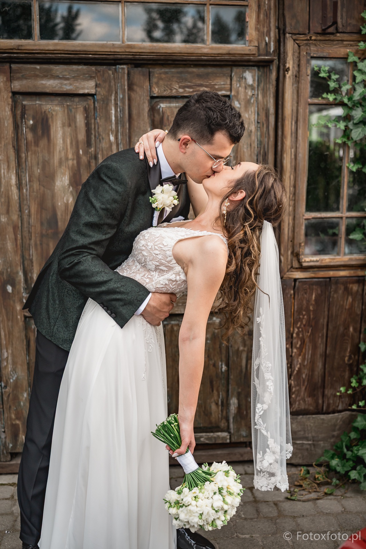 Pocałunek pary młodej przed rustykalnymi, drewnianymi drzwiami z bluszczem. Panna młoda w koronkowej sukni i welonie, pan młody w zielonym smokingu i okularach.