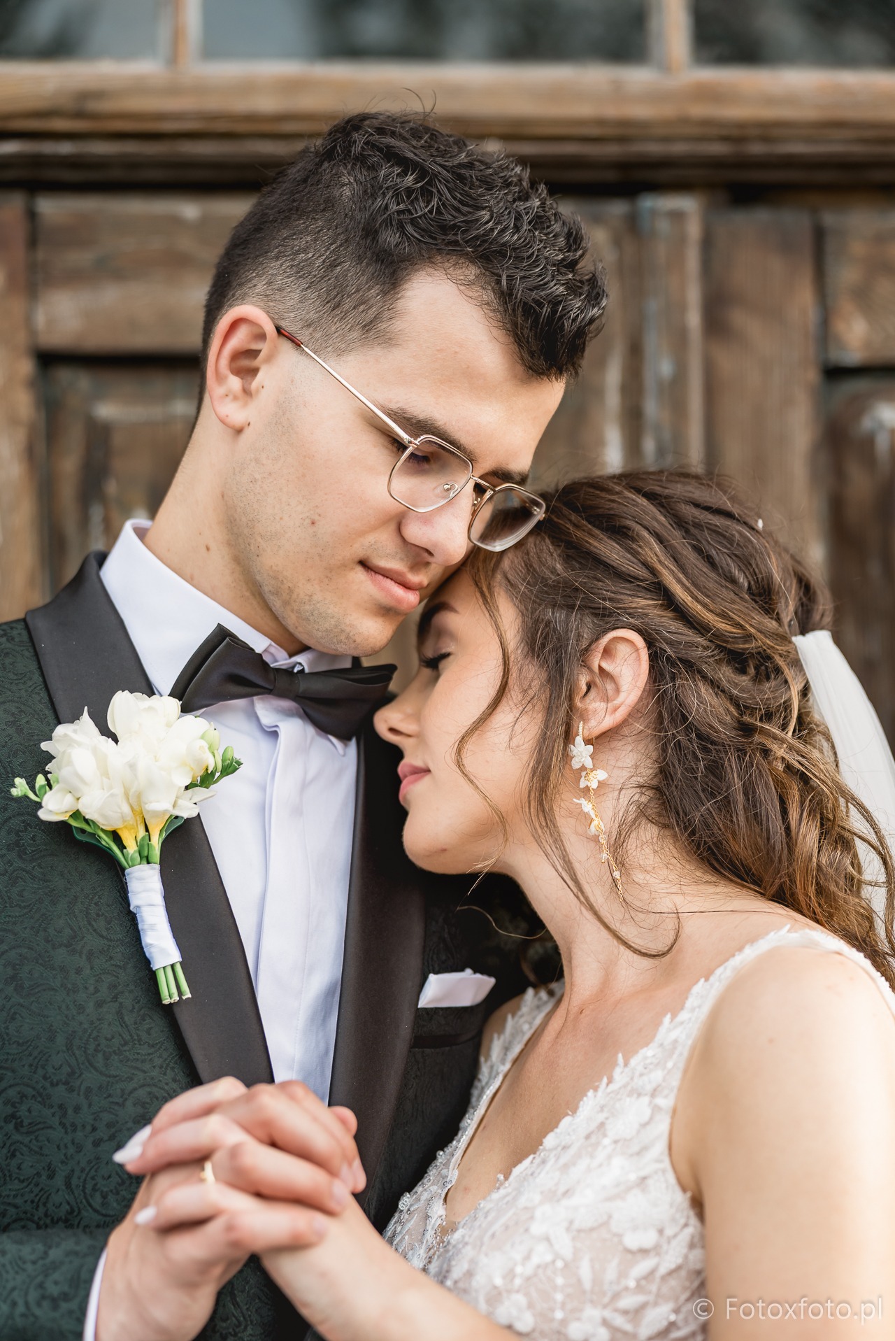 Zamyślona para młoda w zbliżeniu: on w zielonym smokingu z bukiecikiem, ona w białej sukni z koronki i delikatnych kolczykach, na tle drewnianych drzwi.