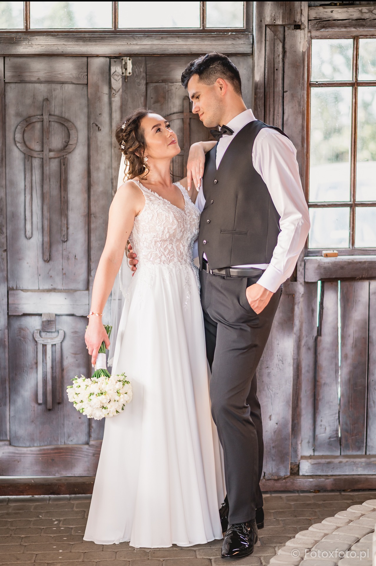 Para młoda w eleganckich strojach pozuje na tle drewnianych drzwi i okna. Panna młoda trzyma bukiet białych kwiatów, a pan młody ma rękę w kieszeni.
