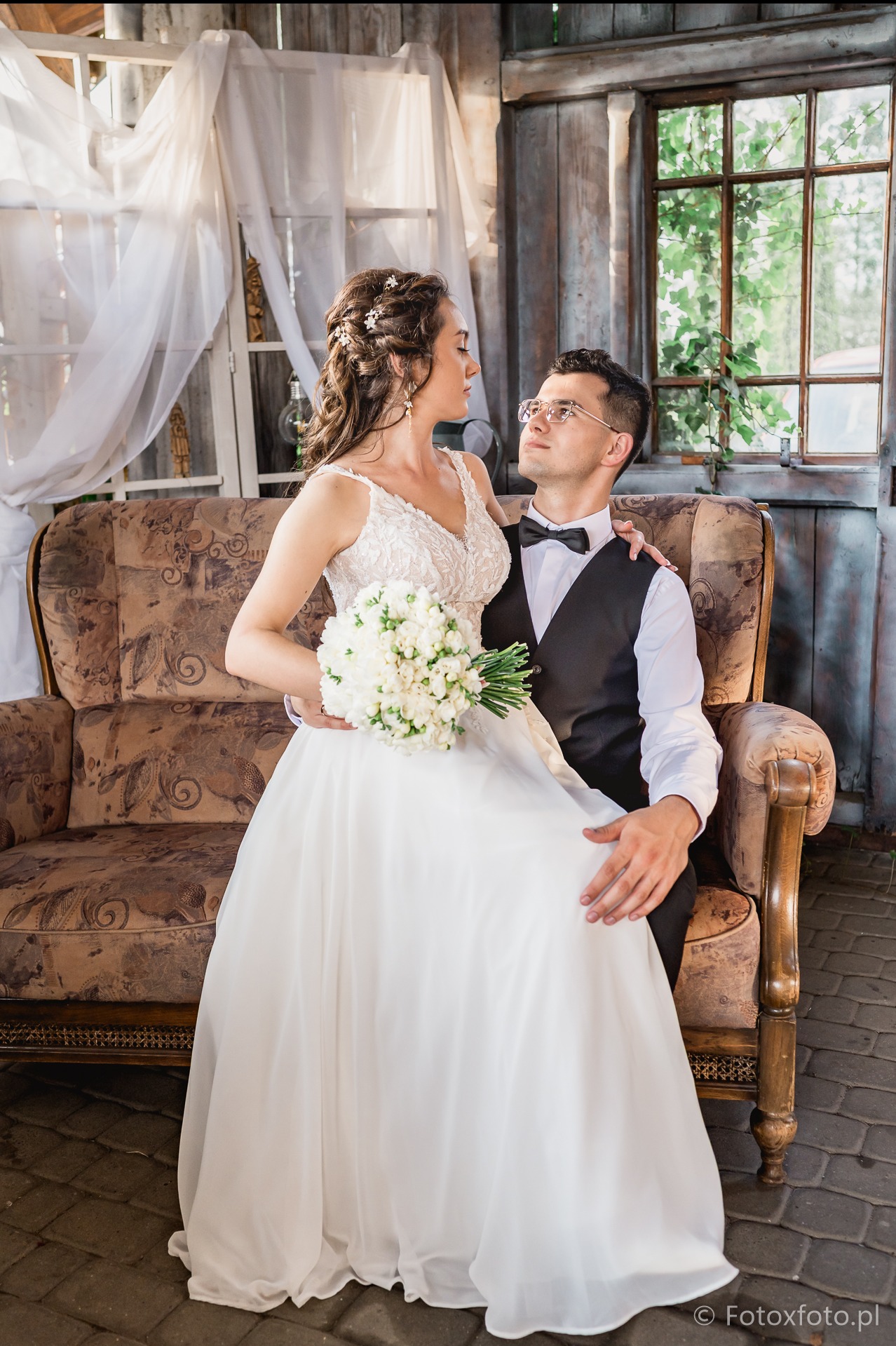 Elegancka para młoda siedzi na zdobionej sofie w rustykalnym wnętrzu. Panna młoda trzyma bukiet białych kwiatów, a pan młody patrzy na nią z czułością.