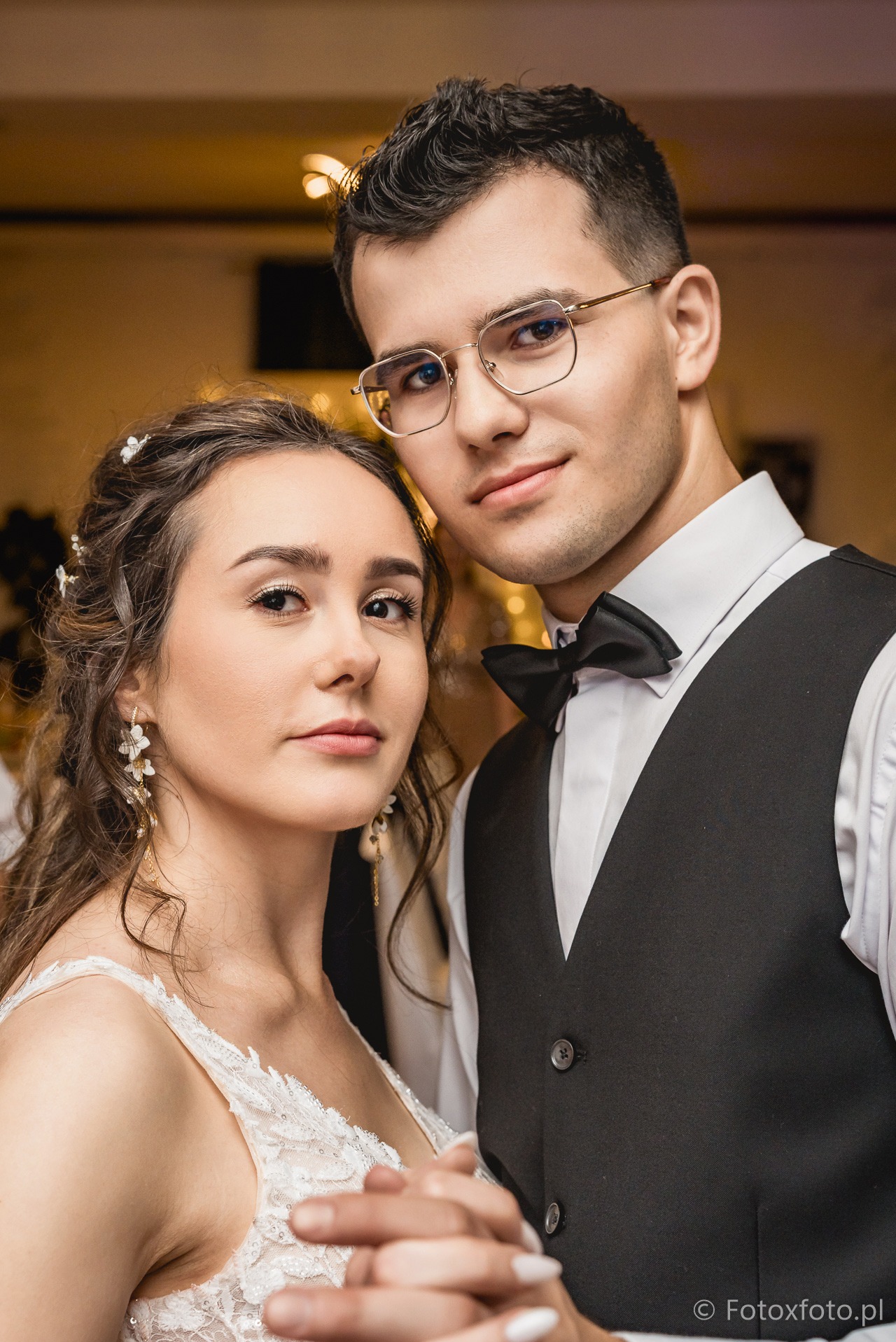 Młoda para w zbliżeniu: ona w białej sukni z koronką i kwiatami we włosach, on w czarnej kamizelce i muszce, oboje trzymają się za ręce, patrząc w obiektyw.