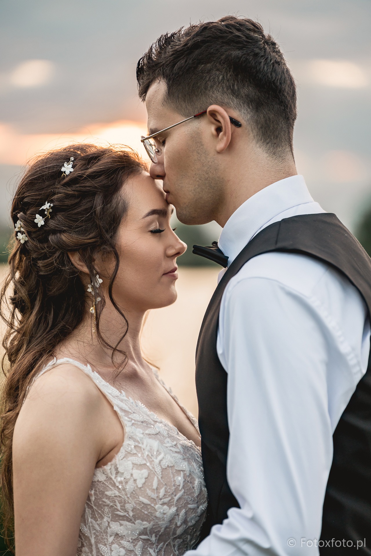 Para młoda w zbliżeniu: on w okularach całuje ją w czoło, ona w białej sukni z kwiatami we włosach, na tle rozmytego krajobrazu i zachodzącego słońca.