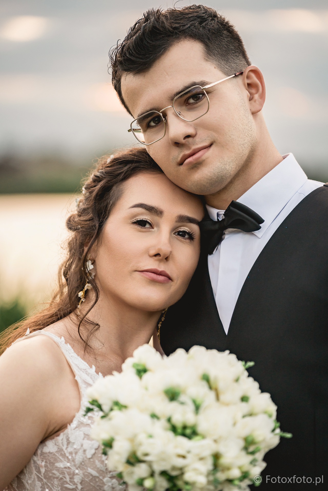 Uśmiechnięta para młoda w zbliżeniu: on w okularach i muszce, ona z bukietem białych kwiatów, subtelne kolczyki, romantyczna atmosfera na tle rozmytej zieleni.