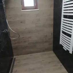 Nowoczesna kabina prysznicowa wykończona płytkami imitującymi drewno i czarny marmur, z grzejnikiem drabinkowym i oknem w tle.