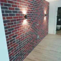 Ściana wykończona dekoracyjną czerwoną cegłą z czarnymi akcentami, oświetlona dwoma nowoczesnymi, czarnymi kinkietami, z widoczną podłogą z paneli imitujących drewno i otwartymi drzwiami do łazienki.