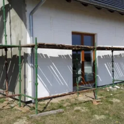 Fragment domu w trakcie ocieplania elewacji, widoczne rusztowania, styropianowe płyty izolacyjne, fragment tynku i okno z brązową ramą.