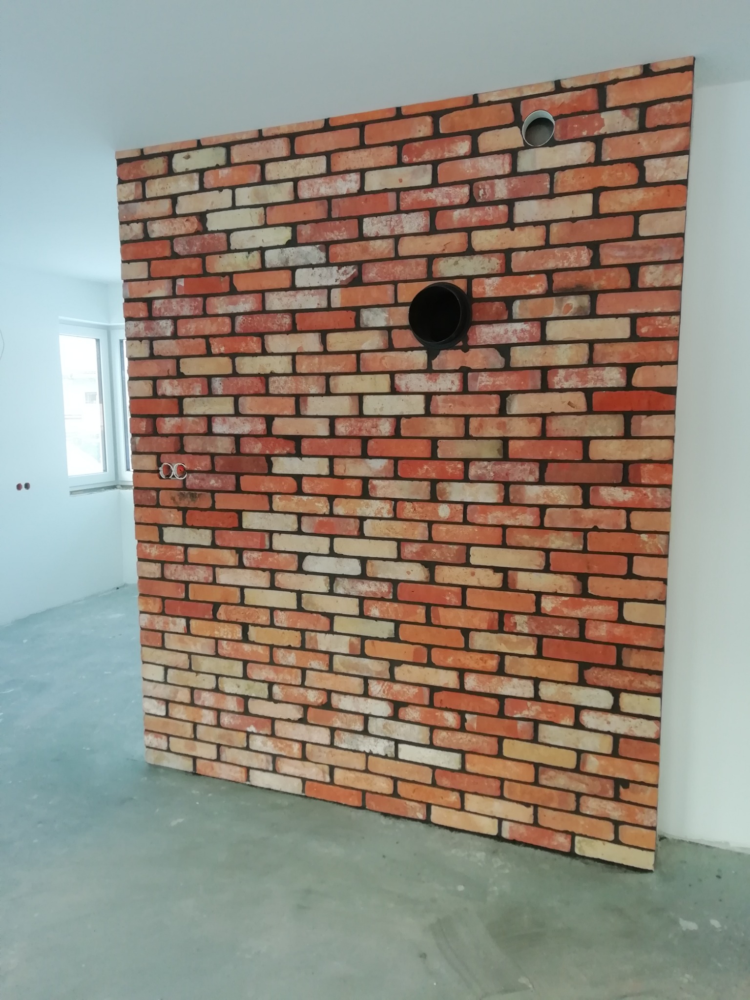 Ścianka działowa z cegły klinkierowej w surowym wnętrzu, widoczne otwory na instalacje, posadzka jastrychowa w tle.