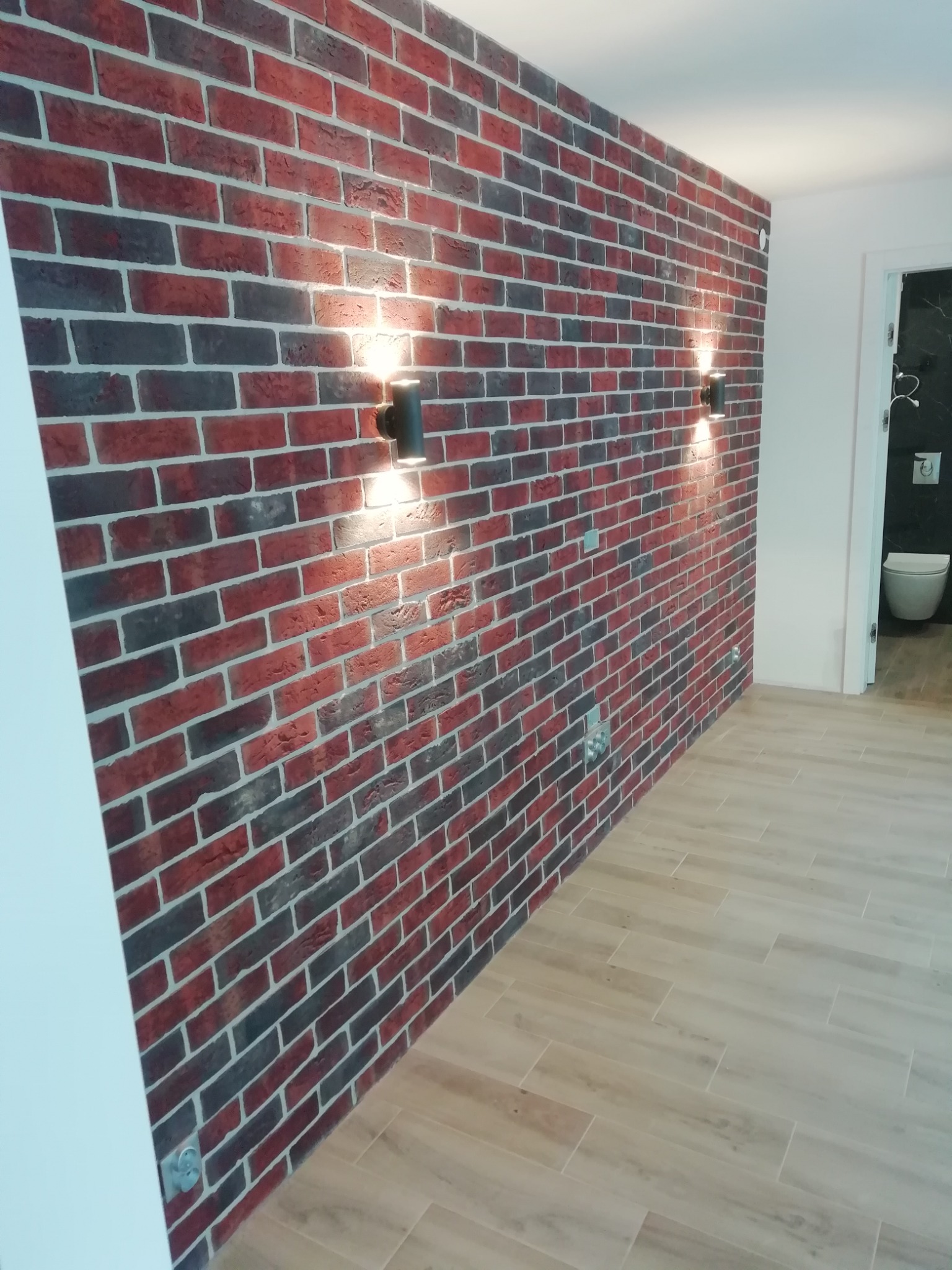 Ściana wykończona dekoracyjną czerwoną cegłą z czarnymi akcentami, oświetlona dwoma nowoczesnymi, czarnymi kinkietami, z widoczną podłogą z paneli imitujących drewno i otwartymi drzwiami do łazienki.