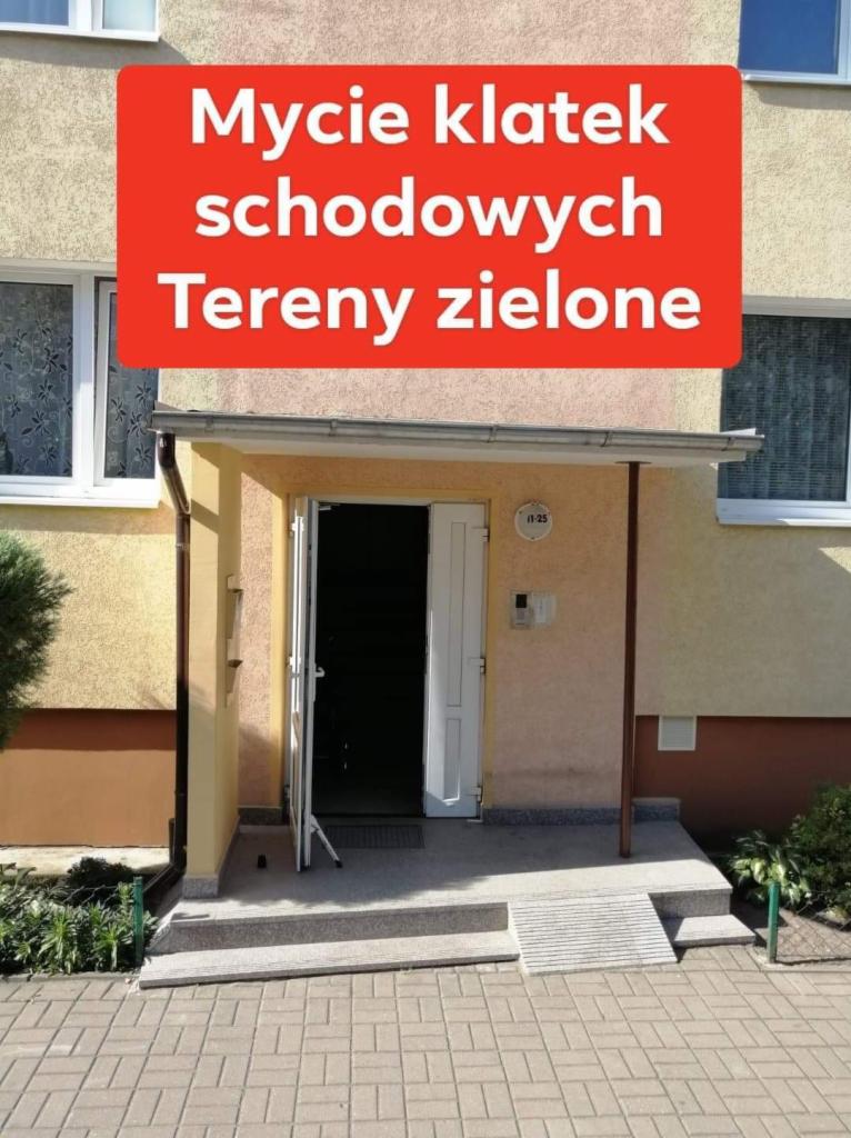 Wejście do budynku z otwartymi drzwiami, widoczna tabliczka z numerem 11-25, chodnik z kostki brukowej i fragment zieleni.