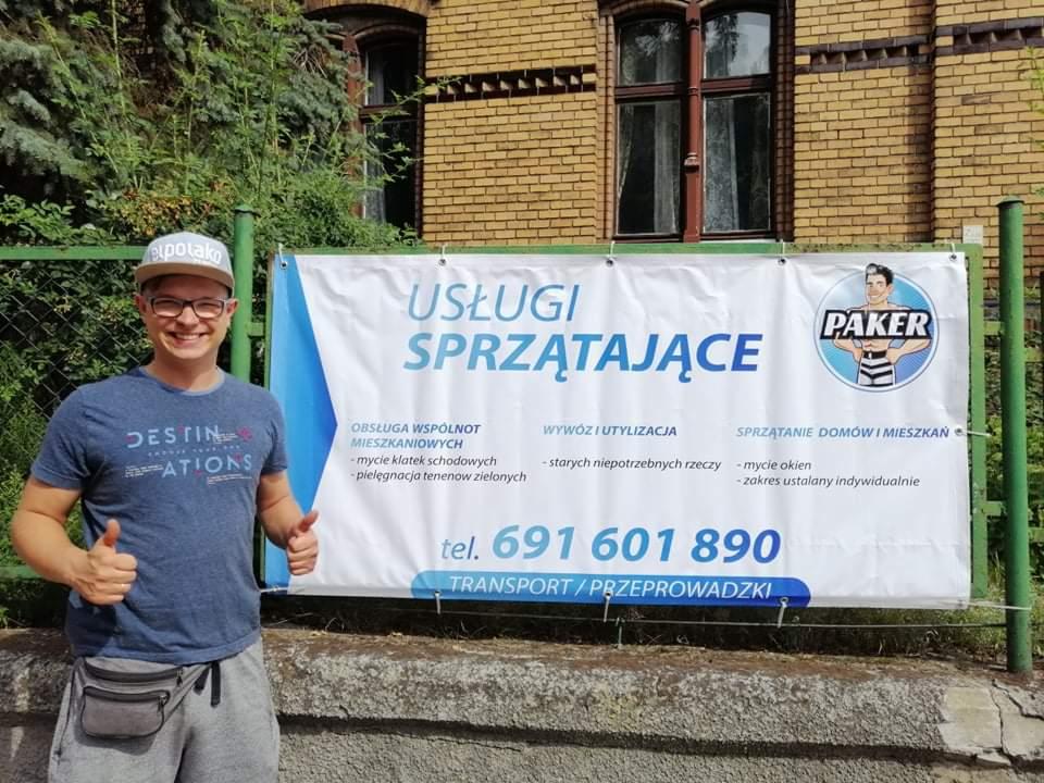 Mężczyzna w okularach i czapce z daszkiem, z uśmiechem i uniesionymi kciukami, pozuje na tle baneru reklamowego firmy oferującej usługi porządkowe, wywóz i transport, z widocznym numerem telefonu.