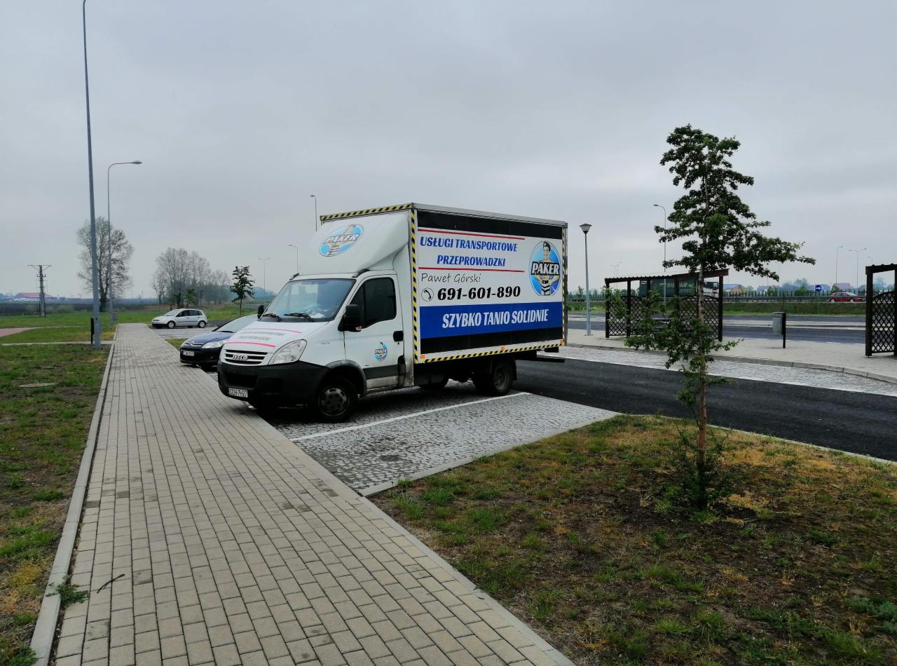 Biała furgonetka firmy transportowej z reklamą usług i numerem telefonu zaparkowana na parkingu z kostki brukowej obok trawnika i młodego drzewa.