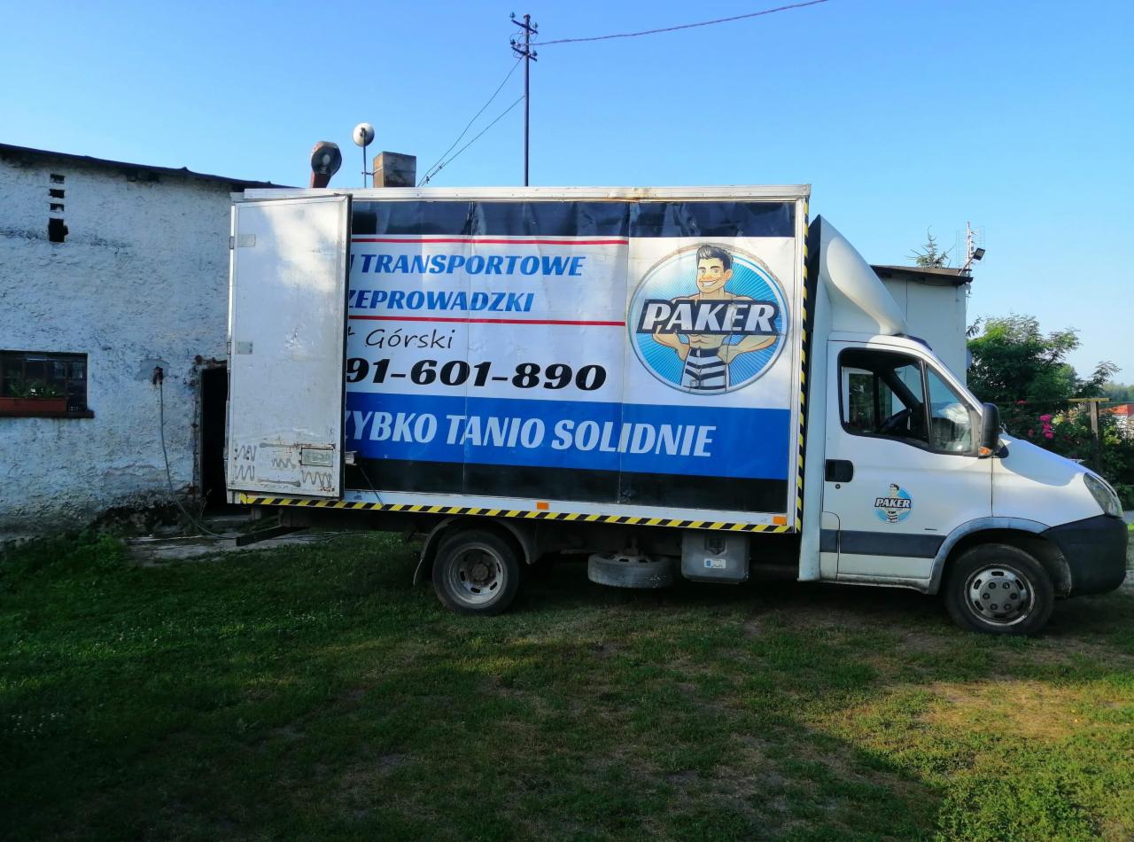 Biała furgonetka z reklamą firmy transportowej 'PAKER' i numerem telefonu zaparkowana na trawniku przed budynkiem.