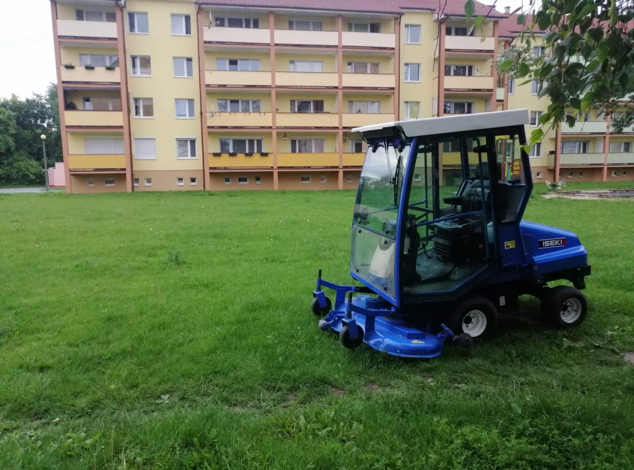 Niebieska kosiarka samojezdna marki Iseki z kabiną, zaparkowana na trawniku przed wielopiętrowym budynkiem mieszkalnym.