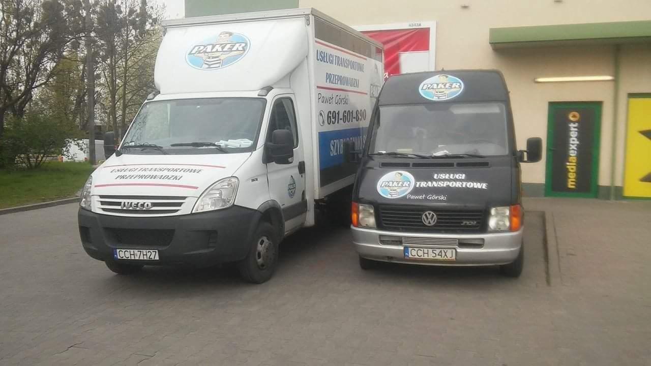 Dwa busy firmy transportowej Paker, jeden biały Iveco z napisem 'Usługi Transportowe Przeprowadzki' i numerem telefonu, drugi czarny Volkswagen, zaparkowane przed budynkiem Media Expert.