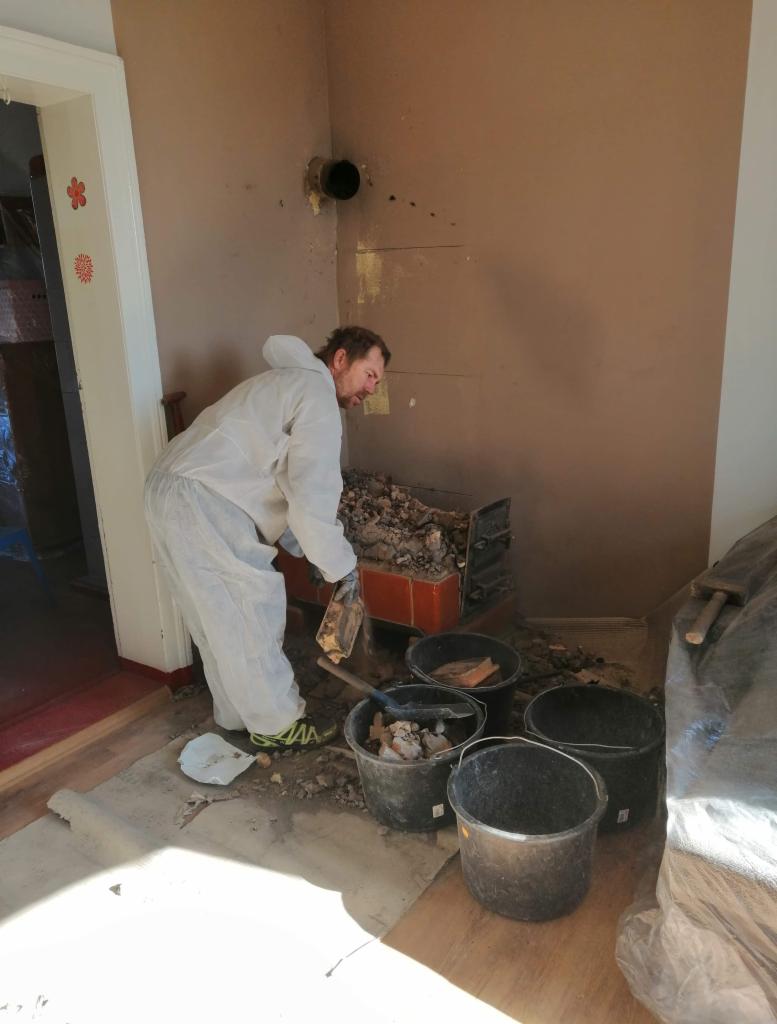 Pracownik w kombinezonie ochronnym usuwa gruz z rozbiórki pieca do czarnych wiader, widoczne ślady po piecu na ścianie i podłodze