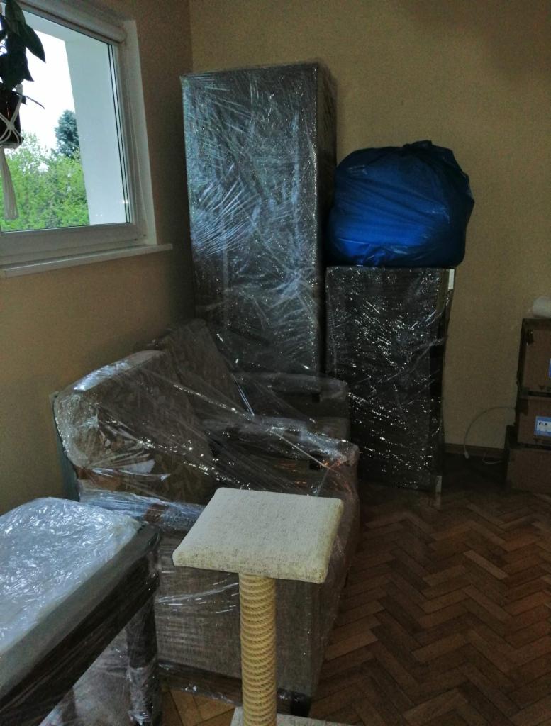 Meble owinięte folią stretch przygotowane do przeprowadzki, widoczny drapak dla kota z jasnym siedziskiem i owiniętym sznurkiem sizalowym słupkiem, parkiet ułożony w jodełkę.