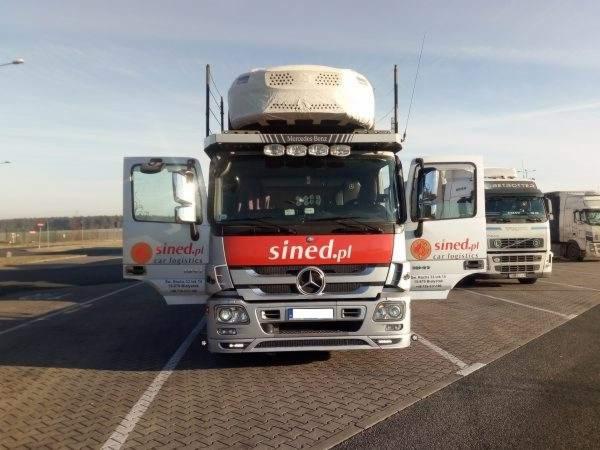 Ciężarówka Mercedes Benz z otwartymi drzwiami, z logo firmy Sined.pl car logistics, zaparkowana na placu z innymi ciężarówkami w tle.