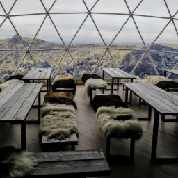 Wnętrze kopuły geodezyjnej z drewnianymi stołami i ławkami pokrytymi islandzkimi skórami owczymi, widok na wulkaniczny krajobraz na zewnątrz.