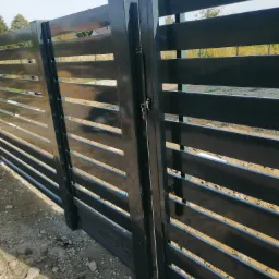 Nowoczesna, czarna brama wjazdowa z poziomych profili stalowych, z widocznym zamkiem i kluczami, zamontowana na żwirowym podłożu.