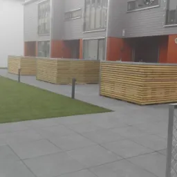 Nowoczesne szeregówki z szarymi elewacjami, drewnianymi osłonami tarasów i szarym brukowanym chodnikiem, z fragmentem trawnika i dekoracyjnymi kamieniami.