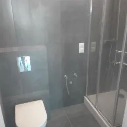 Nowoczesna łazienka z szarymi płytkami, wiszącą toaletą i kabiną prysznicową z widocznymi zaciekami po wodzie.