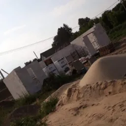 Wznoszone ściany domu z betonu komórkowego, palety z bloczkami SOLBET oraz hałda piasku na placu budowy w słoneczny dzień.