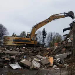 Żółta koparka na tle rozbieranego budynku, z gruzem u podstawy i drzewami w tle. W tle widać również budynek z logo Oferteo.pl