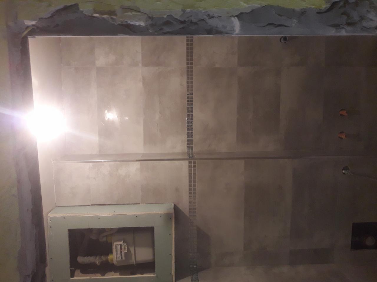 Łazienka w trakcie wykańczania: szare płytki na ścianie, widoczna instalacja podtynkowa WC w obudowie z płyt gipsowo-kartonowych, brak fugi między płytkami, ściana nad płytkami nie wykończona.