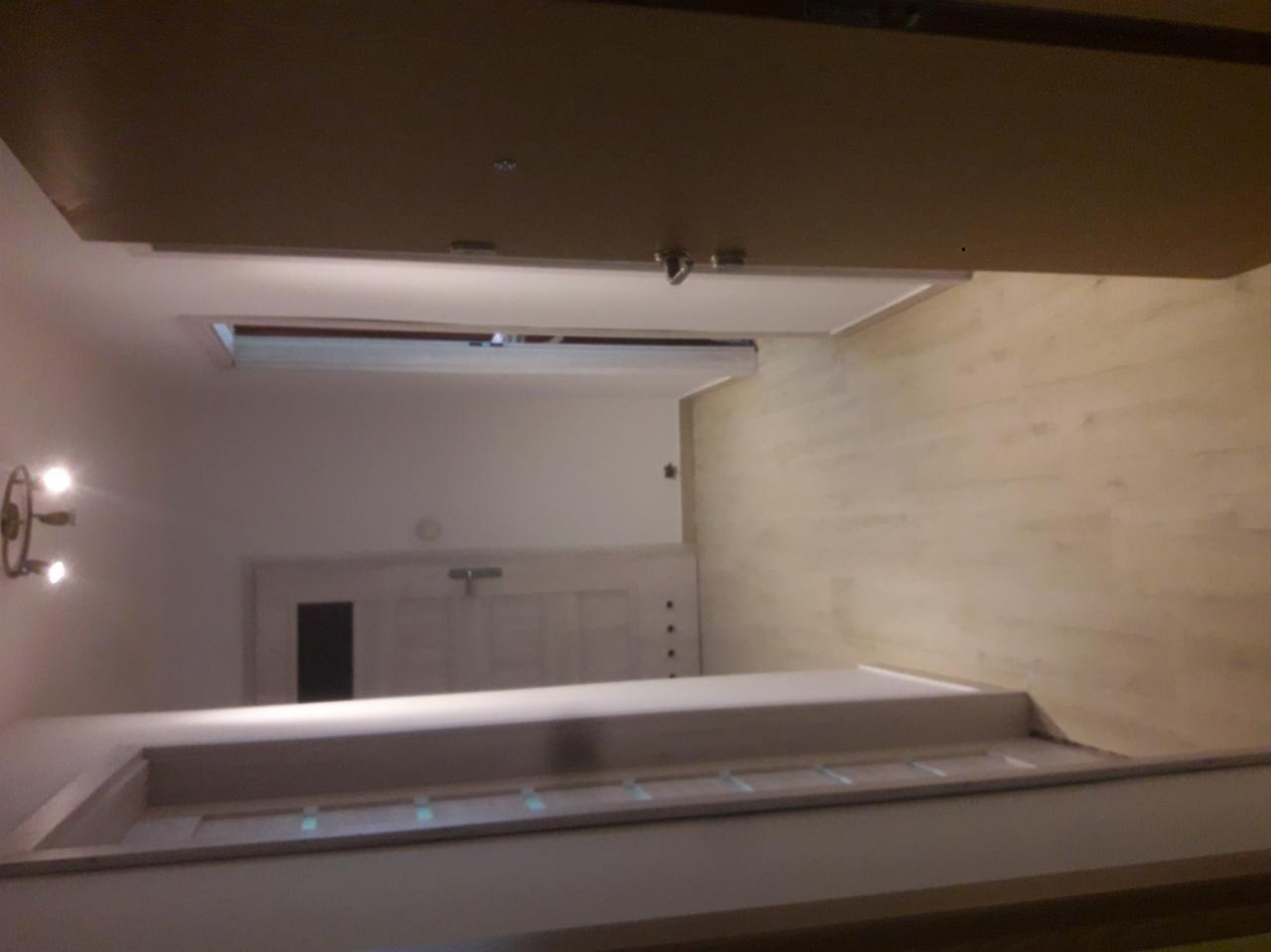 Widok z góry na wykończony korytarz z drewnianą podłogą, białymi ścianami i drzwiami, oświetlony kinkietem z trzema żarówkami.