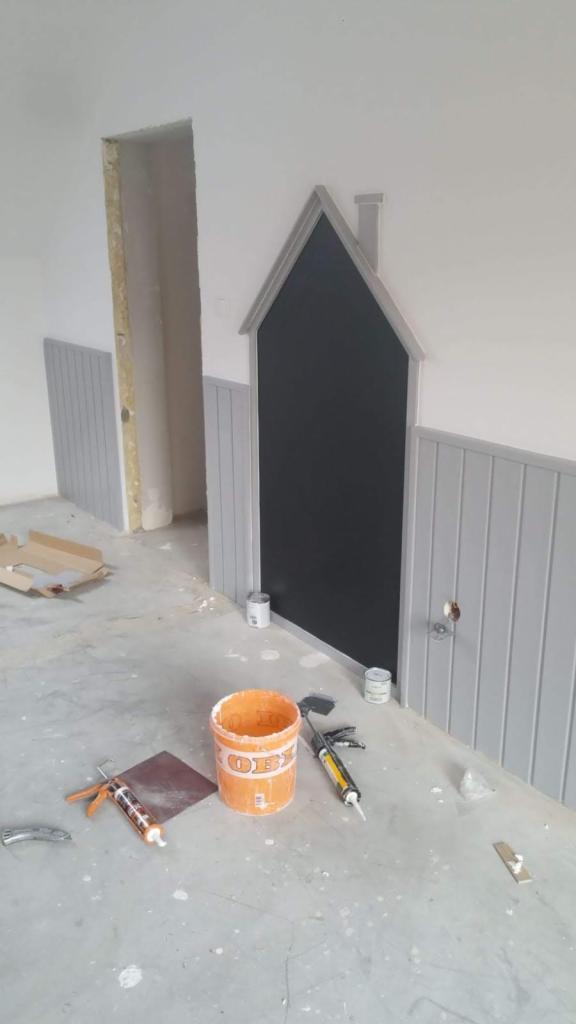 Ściana z szarą boazerią i czarną tablicą w kształcie domku z kominem, podczas prac wykończeniowych, widoczne narzędzia i puszki z farbą na podłodze.