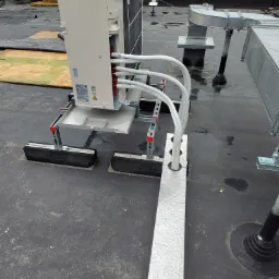 Biała jednostka zewnętrzna klimatyzacji zamontowana na dachu budynku, widoczne rury odprowadzające skropliny i kanały wentylacyjne, mokra nawierzchnia dachu.