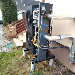 Czarna pompa ciepła typu monoblok na zewnątrz budynku, podłączona do przewodów instalacyjnych, z podłączonym miernikiem cyfrowym podczas serwisu, widoczne elementy wewnętrzne urządzenia.