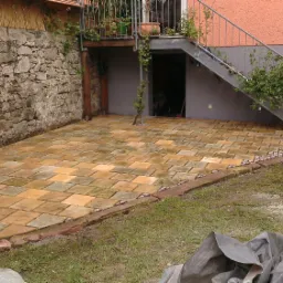 Wyłożony kostką taras z ceglanym obrzeżem, przylegający do kamiennej ściany i budynku z zewnętrznymi schodami, widoczne narzędzia ogrodowe i zwinięty wąż.